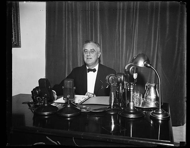 black and white image of FDR sitting at a table with microphones in front of him